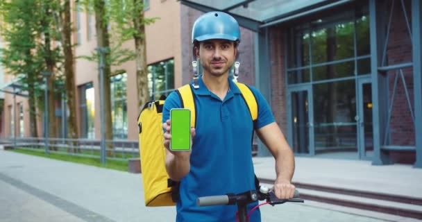 Charmant livreur caucasien avec scooter de pied tenant smarphone avec écran vert. Les jeunes font messager dans le casque de sécurité montrant la clé chromatique sur le téléphone portable. Technologie, concept de service. — Video