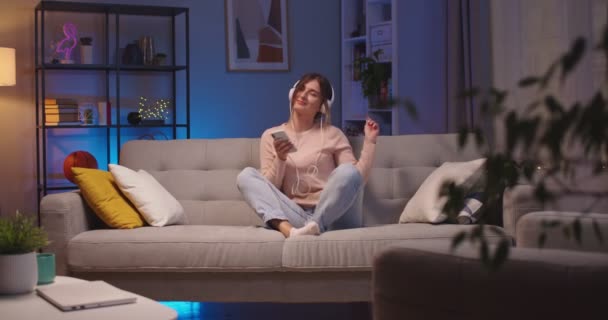 Joyful beautiful Caucasian young woman tapping on smartphone and listening to music in headphones while resting on sofa at home. Happy female enjoying songs from cellphone in room. Leisure concept — Stock Video