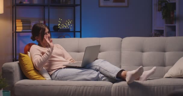 Καυκάσια χαρούμενη όμορφη γυναίκα που έχει βιντεοκλήση στο laptop ενώ ξεκουράζεται στον καναπέ στο σαλόνι σε εσωτερικούς χώρους. Αρκετά χαρούμενη νεαρή γυναίκα βιντεοσκοπεί στον υπολογιστή στο σπίτι της το βράδυ. Έννοια ελεύθερου χρόνου — Αρχείο Βίντεο
