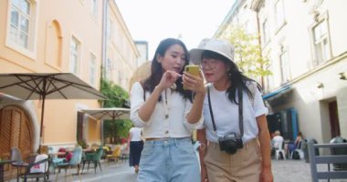 Neşeli Asyalı anne ve kızının birlikte seyahat ettiği portre. Şapkalı güzel kadın gezgin. Genç bayan turist akıllı telefondan bakınıyor ve şehirde yürürken annesine gösteriyor. Turizm konsepti