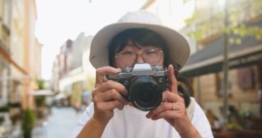 Şapkalı, gözlüklü, sokakta dikilirken fotoğraf çeken neşeli Asyalı güzel bir kadının portresini yakından çek. Neşeli güzel kadın turist dışarıda fotoğraf çekiyor. Kentsel turizm konsepti