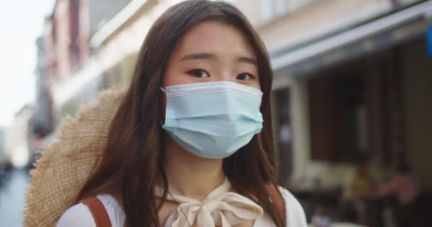 Close up of beautiful young female tourist in mask standing on street and looking away. Pretty Asian woman with calm face in city after quarantine. Protective measures. Safe tourism. Portrait concept — Stock Video