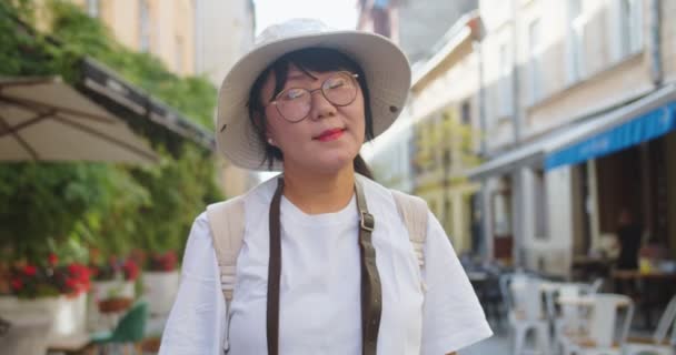 Nahaufnahme Porträt einer fröhlichen asiatischen Reisenden mittleren Alters mit Brille und Hut, die draußen steht und in die Kamera lächelt. Schöne fröhliche Frau in der Stadt auf der Straße. Städtisches Tourismuskonzept — Stockvideo