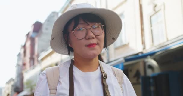 Ritratto di felice turista asiatica di mezza età in occhiali e cappello in piedi in città all'aperto e sorridente. Bella donna gioiosa in città in strada nella giornata di sole. Turismo estivo. Concetto ravvicinato — Video Stock