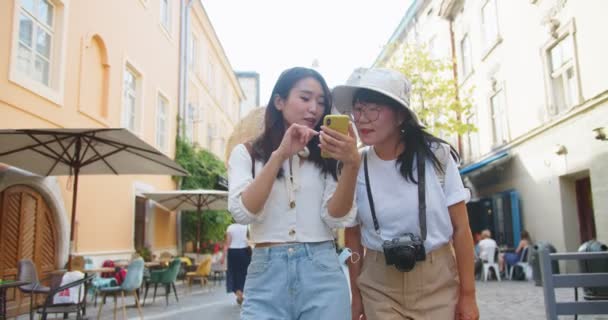 Neşeli Asyalı anne ve kızının birlikte seyahat ettiği portre. Şapkalı güzel kadın gezgin. Genç bayan turist akıllı telefondan bakınıyor ve şehirde yürürken annesine gösteriyor. Turizm konsepti — Stok video