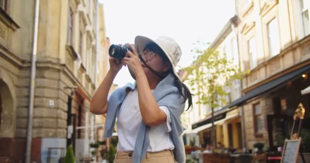 Alegre asiático hermosa hembra en sombrero y gafas de tomar fotos en la cámara mientras camina en la ciudad en la calle. Retrato de mujer bonita feliz toma fotos al aire libre mientras viaja. Concepto de turismo urbano — Vídeo de stock