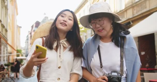 Retrato de la madre y la hija asiática feliz haciendo foto selfie en el teléfono inteligente en la calle de buen humor. Alegre mujer turistas posando mientras toma fotos en el teléfono móvil en la ciudad. Concepto turístico — Vídeos de Stock