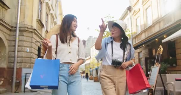 Vacker asiatisk kvinna i hatt talar med ung kvinna när du går utomhus i gamla stan med shoppingväskor. Glädjande mamma och dotter chattar på gatan på gott humör med inköp. Resor shopping — Stockvideo
