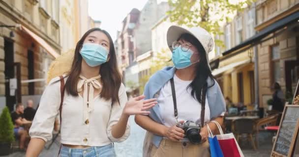 Портрет красивых счастливых азиатских женщин в масках, разговаривающих во время прогулки по старому городу с сумками для покупок. Радостная юная туристка болтает с матерью. Женщина-путешественница с камерой в городе — стоковое видео