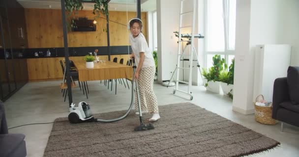 Mujer asiática escuchando música en auriculares blancos que fingen tocar la guitarra mientras limpian al vacío el piso de la alfombra en la sala de estar en casa. Concepto de limpieza, casa de limpieza familiar, estilo de vida. — Vídeo de stock