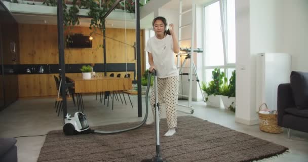 Vidám fiatal, gyönyörű ázsiai nő zenét hallgat fejtámlákban és táncol a modern porszívó szőnyeg padló otthon. A háztartás fogalma, családi takarítás, életmód. — Stock videók