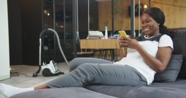 İnternette temizlik yaptıktan sonra kanepeye uzanan neşeli Afro-Amerikalı kadın komik haberler okuyor, göz gezdiriyor, akıllı telefon kullanıyor. Oda servisi, temizlik, yaşam tarzı kavramı..