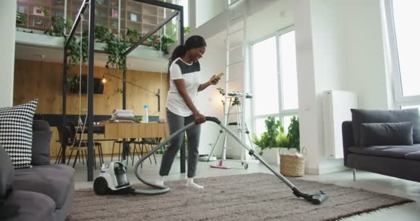 Feliz alegre joven hermosa mujer afroamericana utilizando el suelo de la alfombra de limpieza de vacío moderno en una sala de estar acogedora brillante en casa mientras que hace videollamada con el teléfono inteligente. Concepto de limpieza. — Vídeo de stock