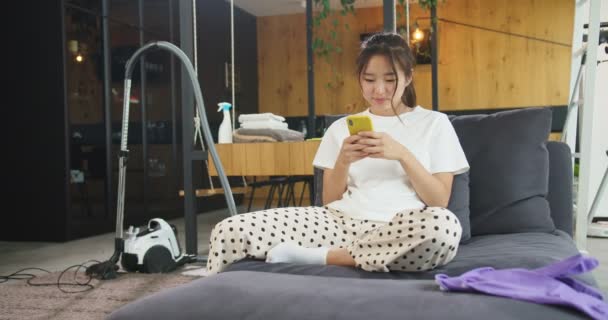 Счастливая веселая молодая красивая азиатка отдыхает на диване после уборки и поиска в Интернете с помощью смартфона. Концепция домашнего хозяйства, уборки дома, образа жизни. — стоковое видео