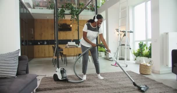 Mujer afroamericana alegre hoovering piso en casa moderna y disfrutar de la música rock en los auriculares cantando y bailando con aspiradora fingiendo tocar en la guitarra. Concepto de limpieza. — Vídeo de stock