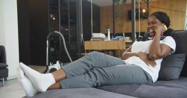 Arkadaşlarıyla konuştuktan ve temizledikten sonra kanepede dinlenen yorgun ve yorgun Afrikalı Amerikalı kadın, dinleniyor, gülümsüyor, akıllı telefon kullanıyor. Oda servisi, temizlik, yaşam tarzı kavramı.. — Stok video