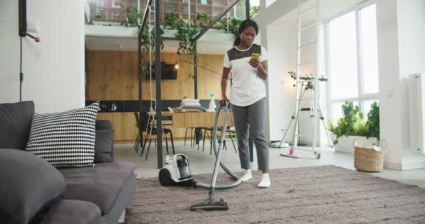 Mujer afro-americana alegre hoovering piso en casa moderna con aspiradora y mensajes de texto de chat con amigos en las redes sociales utilizando el teléfono inteligente. Concepto de limpieza, casa de limpieza, estilo de vida. — Vídeo de stock
