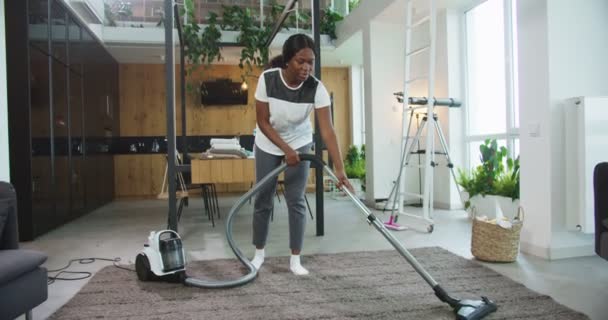 Feliz alegre joven hermosa mujer afroamericana utilizando el suelo de la alfombra de limpieza de vacío moderno en una sala de estar acogedora brillante en casa. Concepto de limpieza, casa de limpieza, estilo de vida. — Vídeos de Stock