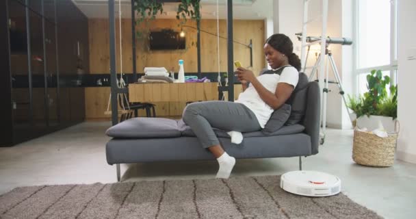 Iloinen afrikkalainen amerikkalainen nainen lepää sohvalla puhdistuksen jälkeen. Nainen, joka nauttii nykyaikaisesta tekniikasta älypuhelimen ja robottipölynimurin avulla, istuu sohvalla kotona. Innovaatiot ja sisustus käsite. — kuvapankkivideo