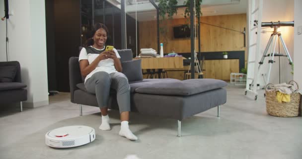 Afro-Amerikalı neşeli genç bayan oturma odasında kanepede oturmuş akıllı telefon kullanarak interneti karıştırırken robotik süpürge de ev işlerini yapıyor. İcatlar ve günlük yaşam konsepti. — Stok video