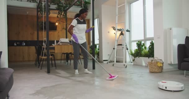 Joyful african american woman mopping floor in living room with mop. Pretty housewife doing housework mopping while robotic hoover cleaning floor. Female concentrated on occupation. Daily routine. — Stock Video