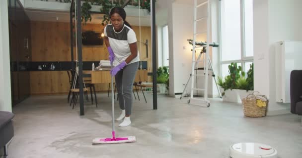 Schöne afrikanisch-amerikanische Frau wischt Boden im Wohnzimmer mit Wischmopp. Hübsche Hausfrau, die Hausarbeit wischt, während Staubsaugerroboter Boden reinigen. Frauen konzentrierten sich auf den Beruf. Tagesablauf. — Stockvideo