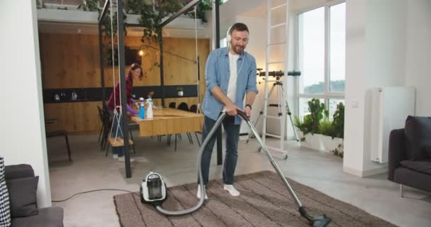 Hombre y mujer feliz pareja de limpieza en casa divirtiéndose. Esposo aspirando piso escuchando música en auriculares y bailando mientras su esposa limpia la mesa. Servicio de limpieza, gente y concepto de felicidad. — Vídeo de stock