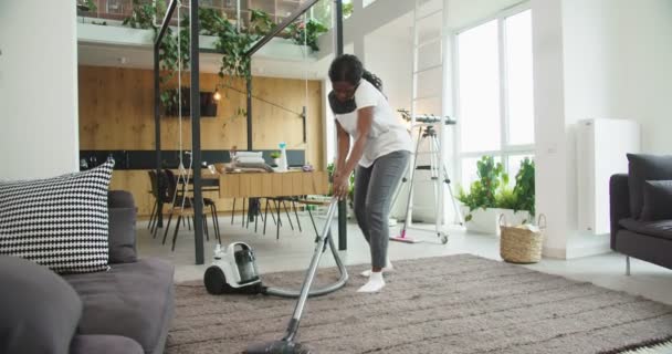 Mujer afroamericana alegre utilizando el suelo de alfombra de limpieza de vacío moderno en una sala de estar acogedora brillante en casa mientras habla por teléfono. Concepto de limpieza, casa de limpieza, estilo de vida. — Vídeo de stock