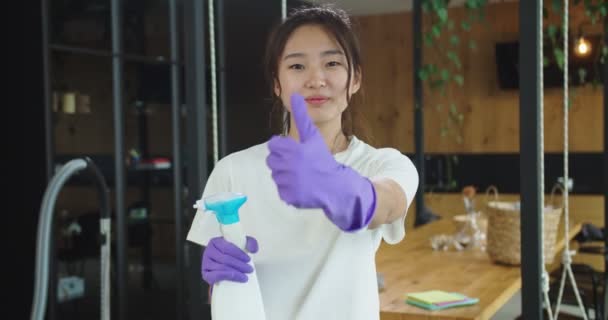 Közelkép egy mosolygó ázsiai háziasszonyról, aki kesztyűs kézzel fogja a tisztább mosószert, és feltartott hüvelykujjakkal és kacsintással néz a kamerába. A háztartás fogalma, családi takarítás, életmód. — Stock videók