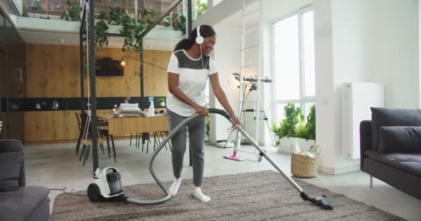 Joyful african american woman hoovering floor in modern house and enjoying music in headphones smiling and dancing with vacuum cleaner. Concept of housekeeping, cleaning house, lifestyle. — Stock Video