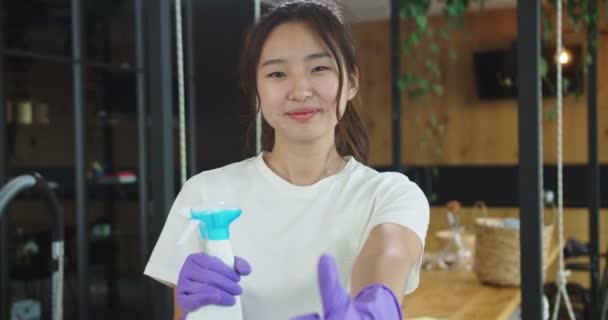 Närbild porträtt av glad leende asiatisk hemmafru hålla renare rengöringsmedel med händerna i handskar och tittar in kameran visar tummen upp. Begreppet städning, familj städhus, livsstil. — Stockvideo