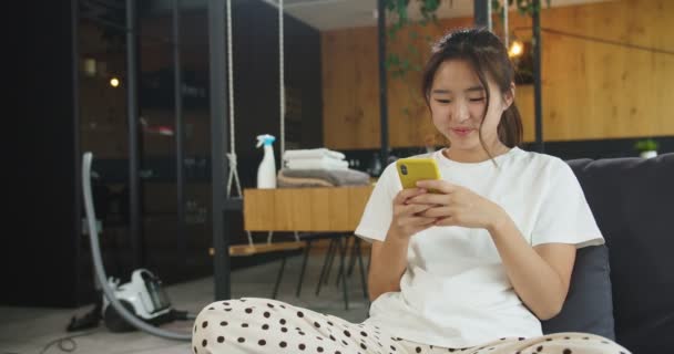 掃除をして彼氏とおしゃべりをした後、ソファの上に寝そべっている幸せな陽気な若いアジアの女性は、スマートフォンを使用してメッセージを書いています。ハウスキーピング、クリーニングハウス、ライフスタイルの概念. — ストック動画