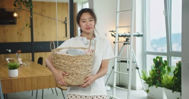Zbliżenie szczęśliwy wesoły młody piękny azjatycki kobieta ze słuchawkami na ramionach trzyma koszyk z brudne ubrania, usługi pralnicze. Koncepcja sprzątania, sprzątania rodziny, stylu życia. — Wideo stockowe