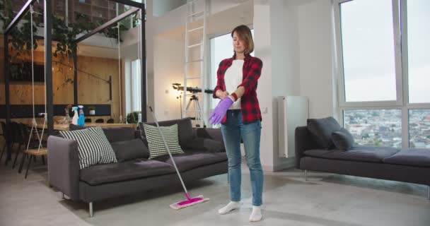 Hermosa mujer en camisa roja que prefigura para la limpieza, usando guantes para fregar el piso en la sala de estar con fregona. Bonita ama de casa haciendo tareas domésticas. Concepto de limpieza, casa de limpieza, estilo de vida. — Vídeo de stock