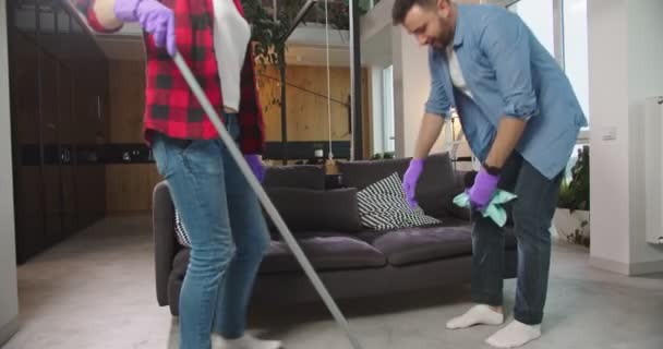 Tired couple relaxing on sofa after clean up house. Wife and husband sitting on sofa in living room taking off gloves and throwing mop. Happy couple cleaning apartment, household, housekeeping concept — Stock Video