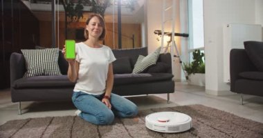 Robotik elektrik süpürgesinin arkasında oturan mutlu bir kadının orta planı. Elinde krom anahtarla telefonu tutması. Yeşil ekranlı telefonu gösteren sevimli bir kadın. Modern teknolojiler, ev idaresi, yaşam tarzı.