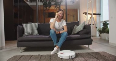 Oturma odasındaki kanepede oturan neşeli genç kadın ev işi yaparken akıllı telefon kullanarak konuşuyor, konuşuyor. İcatlar ve günlük yaşam konsepti.