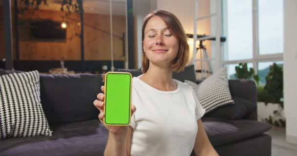 Μεσαίο πλάνο της ευτυχισμένης γυναίκας που κάθεται στο πάτωμα κρατώντας τηλέφωνο με chromakey. Χαριτωμένο θηλυκό δείχνει τηλέφωνο με πράσινη οθόνη. Αντίληψη σύγχρονες τεχνολογίες, chroma κλειδί, smartphone, κινητό τηλέφωνο, τρόπος ζωής. — Αρχείο Βίντεο