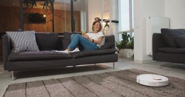 Güzel genç bir kadın robot elektrik süpürgesini açıyor ve otomatik temizliğin tadını çıkarıyor. Kanepede oturuyor ve akıllı telefonuyla dinleniyor. Modern teknoloji ve gençlik kavramı.