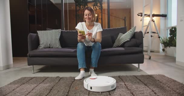 Vidám fiatal nő ül a kanapén a nappaliban böngészi az internetet okostelefonnal, miközben robotporszívózik a padló háztartási munkát végez. Találmányok és a mindennapi élet fogalma. — Stock videók