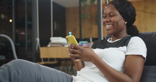 Roupa-se de bela jovem afro-americana dona de casa descansando no sofá após a limpeza e mensagens de texto, bate-papo, leitura, rolagem usando smartphone. Conceito de limpeza, casa de limpeza — Vídeo de Stock