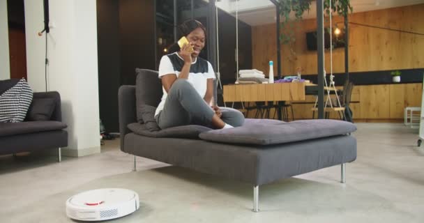 Sonriente joven afroamericana dama sentada en el sofá en la sala de estar hablando llamando usando un teléfono inteligente mientras robótica piso de limpieza de vacío haciendo el trabajo doméstico. Invenciones y concepto de la vida cotidiana. — Vídeos de Stock