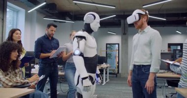 Yeni eserlerini çevreleyen çok ırklı mühendisler ve programcılar. VR kulaklıklı bir erkek hareket ediyor ve insansı robotu çalıştırıyor. Sinerji kontrol ediliyor. Deneme. İnsanlar başarıdan mutlu..