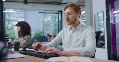 Şık gömlekli ve kızıl saçlı beyaz bir adam yoğun bir ofiste çalışıyor. Klavyede yazıyorum. Birisine kod ya da mesaj yazmak. Daha sonrası için küçük not defterine bir şeyler yazıyorum. Yoğun bir saat. İşyeri.
