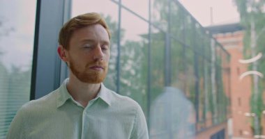 Büyük cam pencerenin önünde duran kızıl saçlı ve sakallı yakışıklı beyaz adam. Modern bina. Erkek, kameraya özgüvenli ve ciddi bir ifadeyle bakıyor. Ofis binası Girişimci.