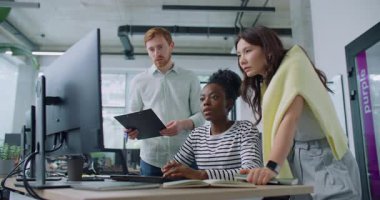 İki farklı etnik gruptan yöneticinin iş arkadaşlarının bilgisayar üzerindeki işlerini tamamlamalarına yardım ettiği bir kamera görüntüsü. Ciddi bir kadın masada oturuyor ve teknoloji cihazına bilgi giriyor. Takım çalışması kavramı.