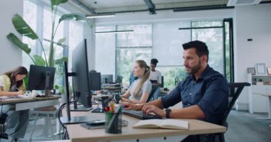 Kısa siyah saçlı ve sakallı meşgul bir adam masada oturuyor ve bilgisayardaki verileri kontrol ediyor. Mavi gömlek giyip ekrana odaklanmış bir şekilde bakıyorsun. Arka planda teknoloji cihazları üzerinde çalışan insanlar.