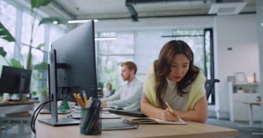 Masada otururken kendine güvenen bir kadının önemli bilgileri not ettiği kamera görüntüsü. Şirket bilgisayarını kullanan çalışkan bir kadın. Arka planda çalışan meşgul yöneticiler. Meslek kavramı.
