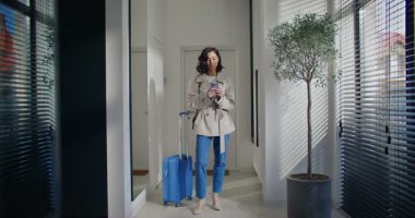 Mutlu beyaz kadın evden ayrılmaya hazır. Mavi bavulun yanında duruyor. Ceket giyiyor. Ayrılmadan önce mobil cihaz kullanılıyor. Metin yazılıyor veya ileti giriliyor. Arkadaşınla ya da ailenle konuş..