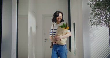 Kameralar beyaz bir kadına doğru yaklaşıyor. Bir çanta dolusu taze yiyecekle lobinin ortasında duran bir kadın. Satın alınmış mallara bakıyorum. Gülümserken doğrudan kameraya bakıyorum..